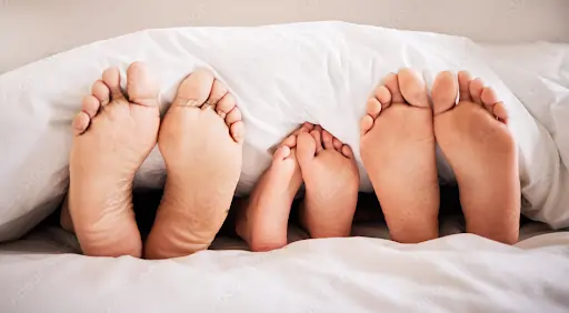 A row of three pairs of feet as seen from the soles, with legs and up covered by a duvet.
